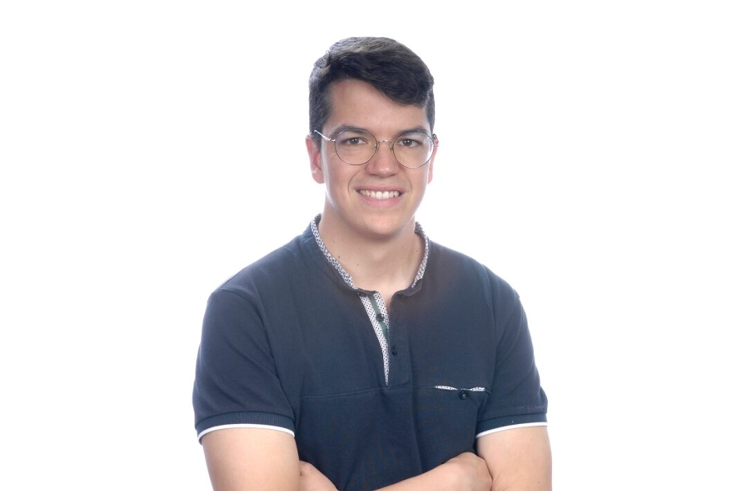 Joven con gafas y cabello corto, sonríe con brazos cruzados, vistiendo camiseta oscura sobre fondo blanco.